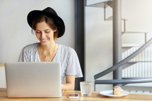 Pretty young female copywriter wearing black hat and stripped blouse working on laptop computer, loo