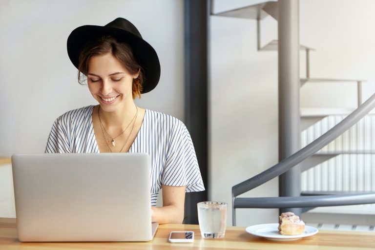 Pretty young female copywriter wearing black hat and stripped blouse working on laptop computer, loo